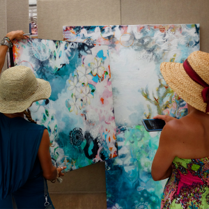 “A photograph of two women looking at 3 paintings, one woman is holding a painting and the other is holding a phone.”